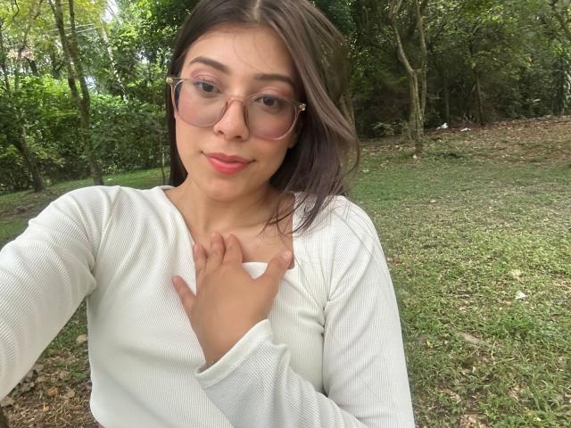 IsabelaPretty with glasses, pink lips, white long-sleeve shirt, hand on chest, green forest background, brown hair, slight smile, light brown skin, medium breasts, casual selfie.