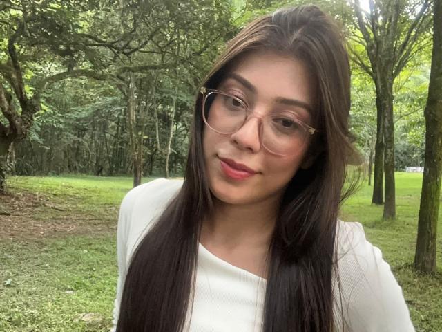 IsabelaPretty with long brown hair, wearing glasses and a white top, has pink lipstick and a slight smile, standing in a green wooded area.