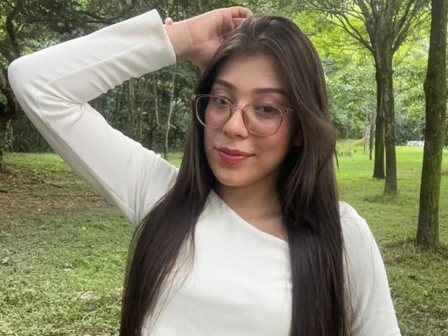 IsabelaPretty with long dark hair, wearing glasses and a white long-sleeve shirt, has a hand in her hair, smiling slightly, showing red lipstick, in a green forest.