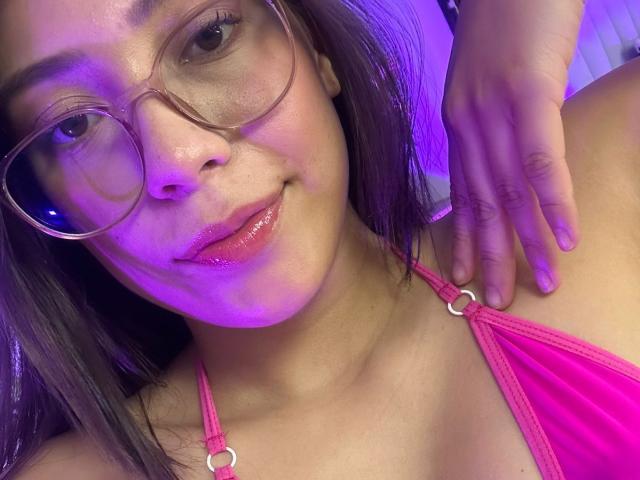 IsabelaPretty with shiny pink lips, wearing clear glasses and a pink halter top, has a hand on her shoulder, showing a slight smile with purple lighting.