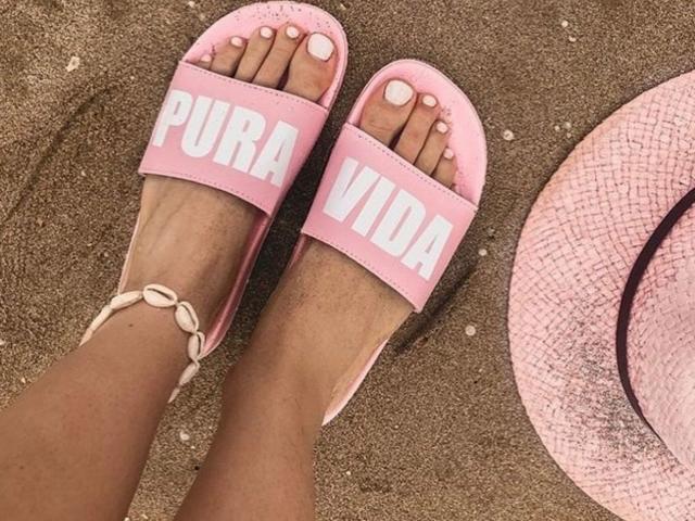 RubyLuna's feet in pink "PURA VIDA" sandals with white letters, white toenails, ankle bracelet, and sandy toes, wearing a pink straw hat on brown sand.