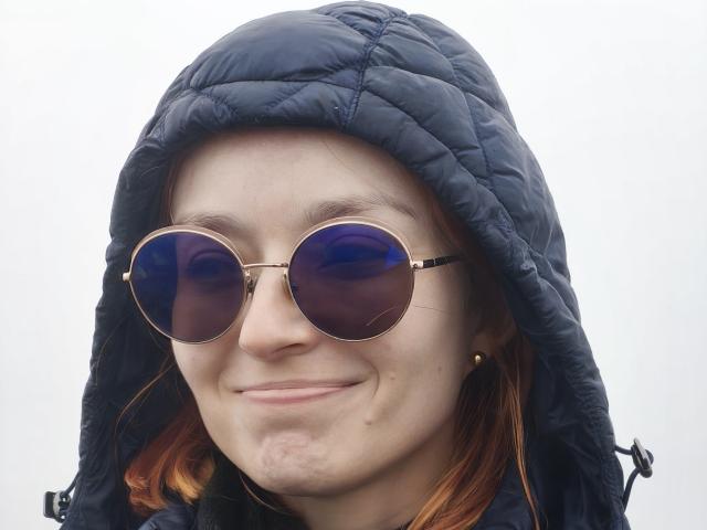 SarahRousen with red hair, wearing a black quilted hood and round sunglasses with blue reflective lenses, smiling slightly. Her face is pale with small acne marks.