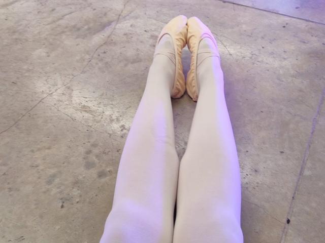 CurlyBlossom's legs in pale tights, standing with toes pointed inward, wearing beige ballet flats, on a textured, concrete floor, showing a relaxed, almost straight, stance.