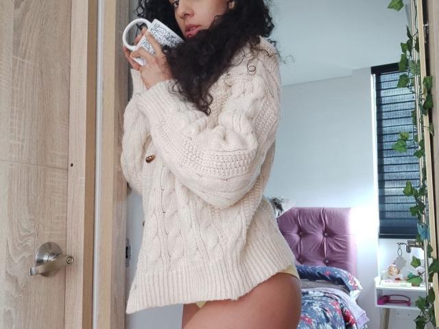 CurlyBlossom with curly black hair, wearing a chunky white knit sweater, holding a white mug, and showing her bare yellow panties and butt. Bedroom setting, daytime.