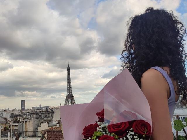 CurlyBlossom with curly black hair, holding a pink bouquet of red roses and white flowers, stands with her back to the Eiffel Tower. Sky is cloudy.