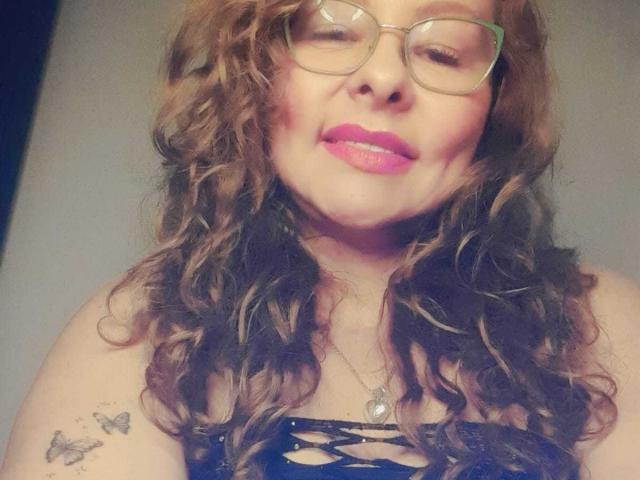 MiaAmoretti with curly brown hair, glasses, pink lipstick, black lace top, butterfly tattoo on left shoulder, smiling softly. Her tits are covered by the lace top.