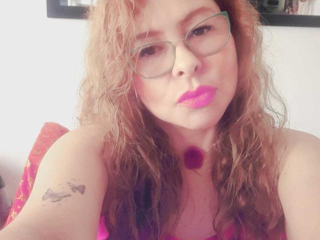MiaAmoretti with wavy brown hair, glasses, pink lipstick, and a red necklace, showing a tattoo of birds on her left arm, looking at the camera. Boobs visible.