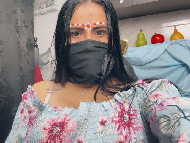 DaliaDubey with long black hair, wearing a floral dress, black mask, and red patterned forehead dot. She has small tits and a tattoo on her left chest.