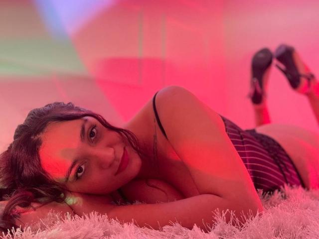 IvyJoness está acostada boca abajo sobre una alfombra blanca y esponjosa, con iluminación roja y rosa, vistiendo una blusa a rayas negra y rosa, y tacones negros, mostrando su trasero..