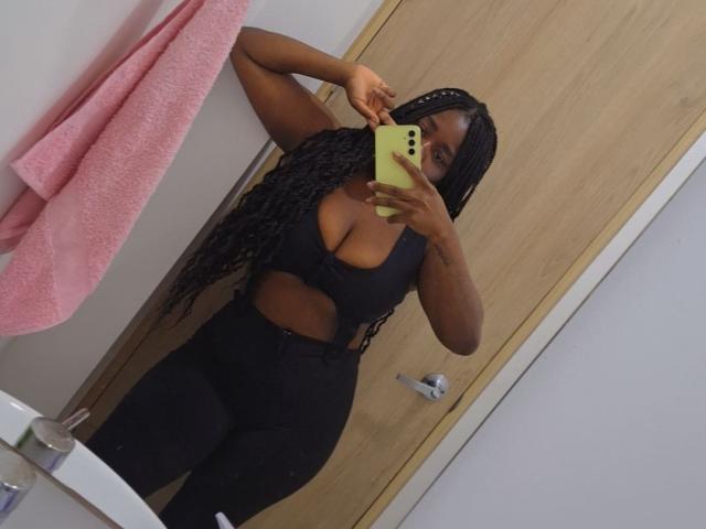 LeidySwet with long braids, black cut-out top showing large tits, black high-waisted pants, taking mirror selfie in bathroom, holding yellow phone, pink towel on wall.