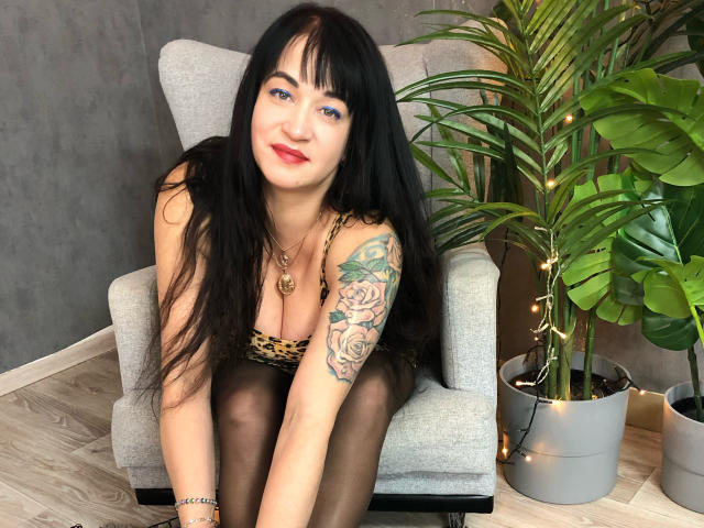 CharlotteMi with long black hair, blue eyes, red lipstick, leopard print top, black tights, tattoo on right arm, sitting in gray chair, potted plant on right, wooden floor.
