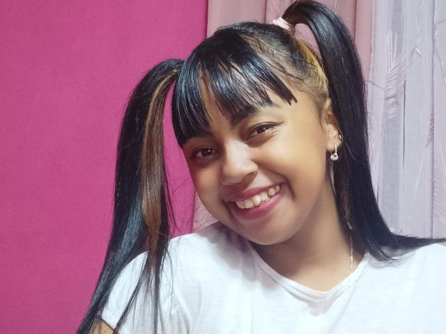 MaliciaQueen with black and brown twin pigtails, smiling, wearing a white shirt, pink and white background, small earrings, light brown skin, and a cute, sexy, happy face.