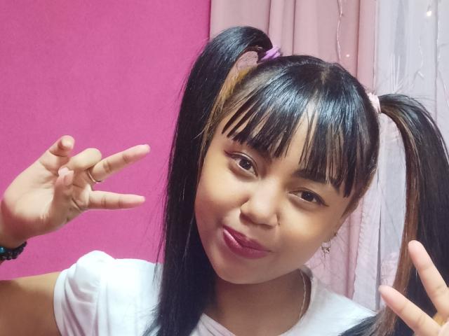 MaliciaQueen with black pigtails, pink lip gloss, and white shirt, making a peace sign with her right hand, smiling slightly. She has a cute, sexy look.