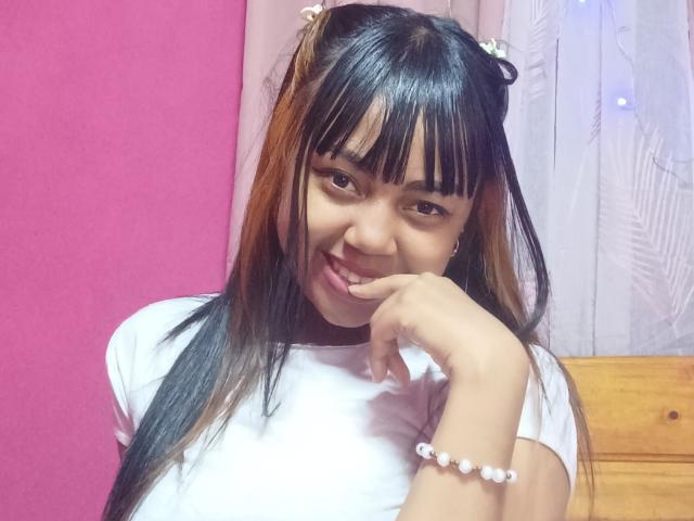 MaliciaQueen has black and brown hair, bangs, and a white shirt. She's smiling, biting her finger, with a pink background and a white curtain. Her ass is not visible.