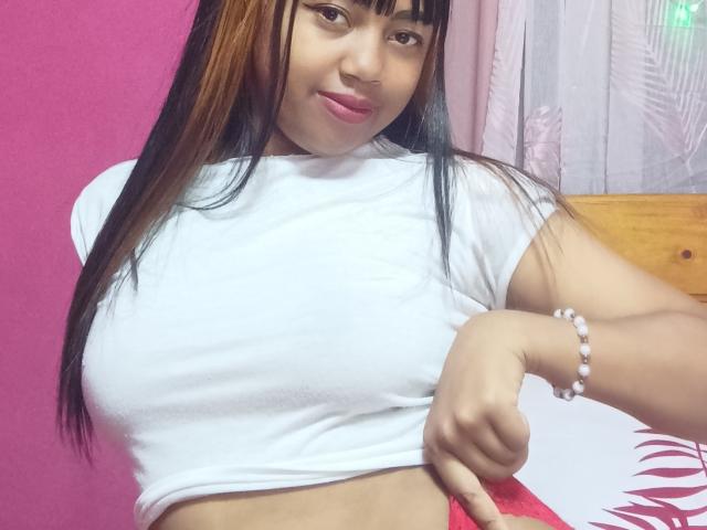 MaliciaQueen with long brown and black hair, white t-shirt, and pink lipstick, showing her tits by lifting her shirt, wearing a white bead bracelet, smiling slightly.