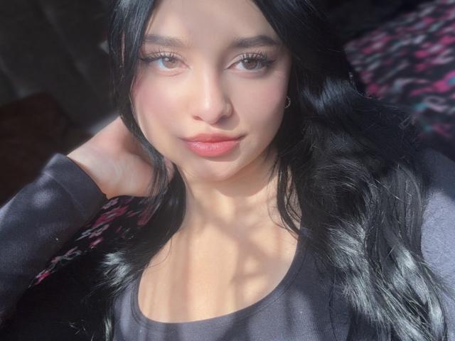MeganPaige with long black hair, light skin, and pink lips, wearing a black shirt, has her hand in her hair, and is looking at the camera with a slight smile.