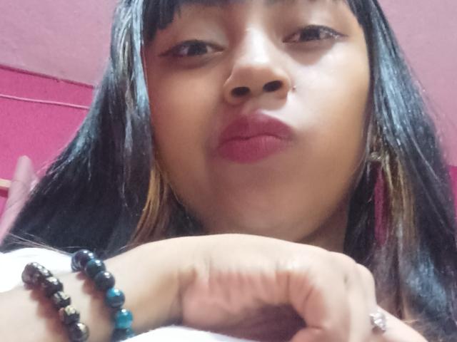 MaliciaQueen has dark brown hair, pink lipstick, and is wearing black and blue beaded bracelets. Her hand rests on her chin, showing a slight pout.
