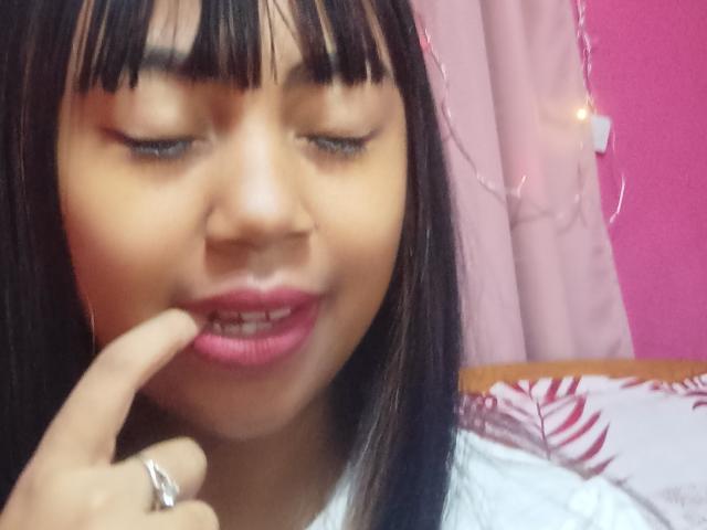 MaliciaQueen with brown hair and pink lipstick, eyes closed, touching her lower lip with her finger, wearing a white shirt, pink curtain, and string lights in the background.