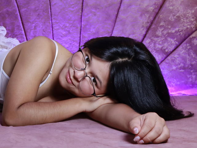 Mei69 with glasses, lying on pink fabric, white lace top, black hair, smiling, resting chin on hand, showing cute, soft, sexy face, and small tits.
