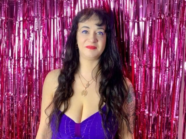 CharlotteMi with long black wavy hair, blue eyes, red lipstick, purple sparkly dress, showing big tits, standing in front of pink metallic streamers, wearing a necklace with a heart.