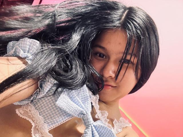 Mei69 with black hair, wearing a blue and white checkered top with lace, has her hair partially covering her face, looking at the camera with a serious expression.