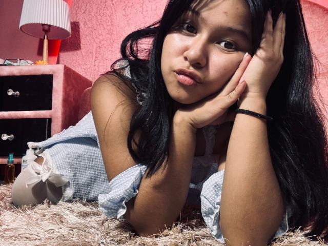 Mei69 with long black hair, pouty lips, and tan skin, resting her head on her hand, wearing a blue checkered dress, on a furry beige rug.