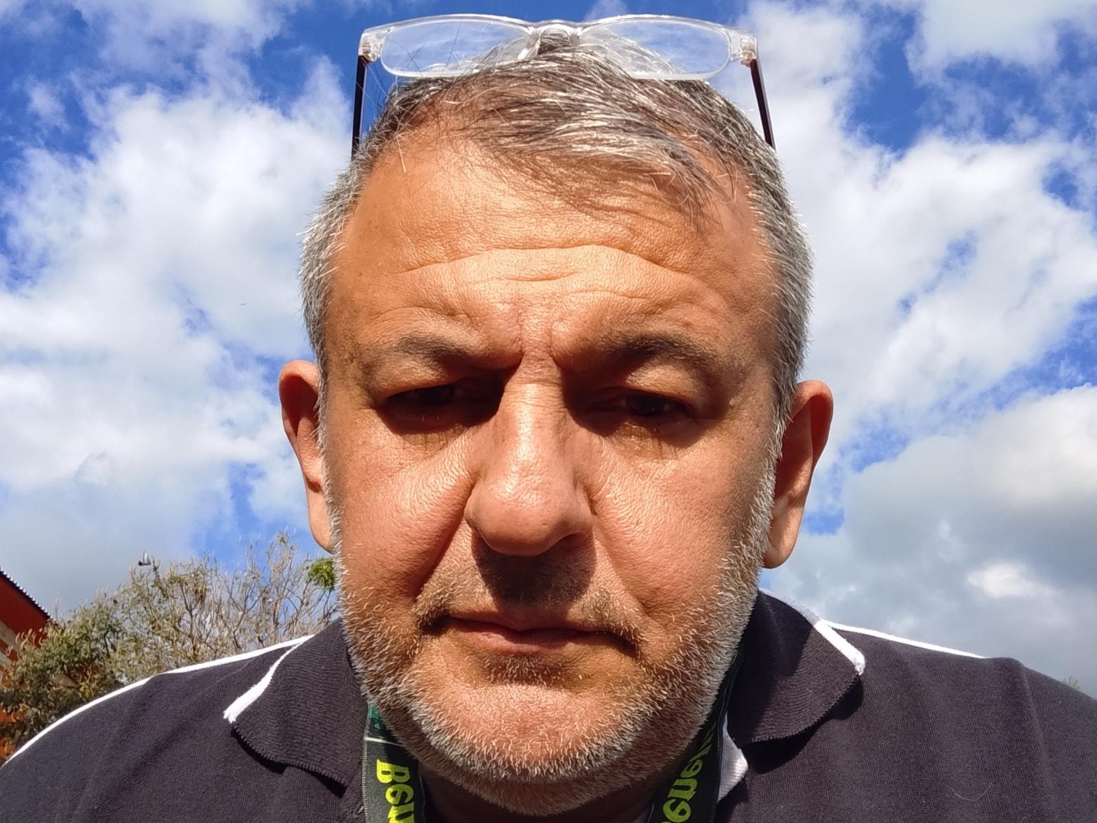 Middle-aged MaxPot with gray hair, beard, and sunglasses on head, looking down with a slightly annoyed expression, wearing a dark polo shirt and green lanyard.