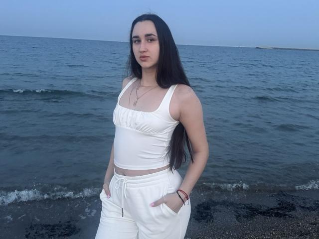 FortuneLisa69 a de longs cheveux noirs, un débardeur blanc et un pantalon blanc, les mains dans les poches, debout près de l'océan, avec un ciel bleu et une eau calme, et elle a l'air sérieuse..