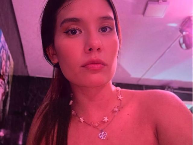 DannaHeat with pink-tinged skin, brown eyes, and long brown hair, wearing a star necklace, looking at the camera with a neutral expression. Boobs are not visible.