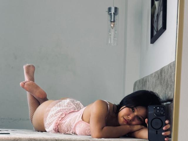Mei69 lag auf einem Bett, trug rosa Spitzendessous und Netzstrümpfe, hielt ein Telefon und hatte ihren Hintern und ihre Brüste sichtbar, während sie in die Kamera blickte..