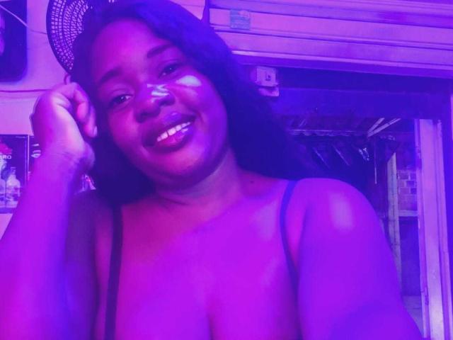 LeidySwet with purple-lit skin, smiling, dark hair, no clothes, showing tits, hand on head, in a room, looking at camera.