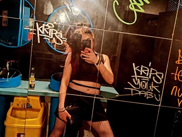 VanessaDomy with long brown hair, wearing a black crop top, fishnet stockings, and a black skirt, taking a mirror selfie in a gritty bathroom, holding a phone.