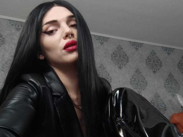 VanessaDomy with long black hair, black leather jacket, red lipstick, nose ring, holding shiny black boots, looking down, pale skin, subtle makeup, dark eyes, slightly open mouth.