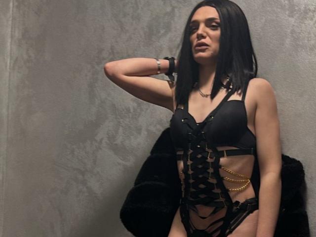 VanessaDomy with black hair, wearing a black leather strappy lingerie set, gold chain, and wrist bracelet, standing against a textured gray wall, hand on head.