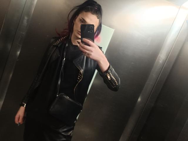 VanessaDomy with black leather jacket, gold studs, black shirt, black pants, and black crossbody bag, taking a mirror selfie with a phone, pink-tipped hair in a ponytail.