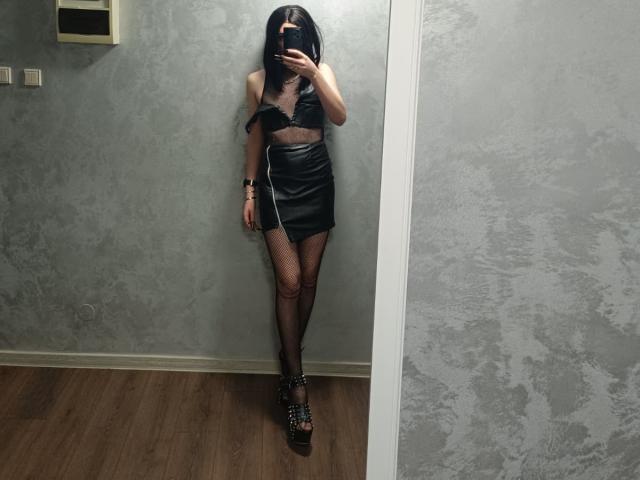 VanessaDomy stands in front of a mirror, taking a selfie. She's wearing a black, strapless, lace-up top with a cutout, and a matching black leather mini skirt. Fishnet stockings cover her legs, and she's wearing black, strappy, platform sandals with metal studs. VanessaDomy's dark hair is straight and falls to her shoulders, partially covering her face as she holds the phone. The room has a textured gray wall and wooden floor. Her right arm is slightly raised, and her left arm is relaxed by her side. The overall style is edgy and provocative.