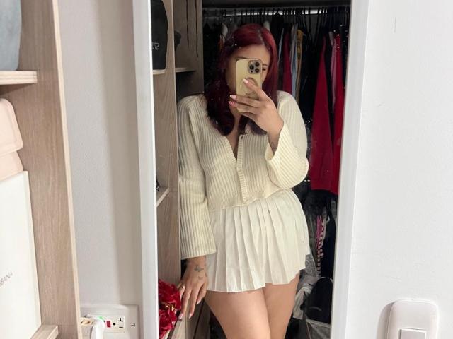 ElyRoss with red hair, wearing a white ribbed sweater and pleated skirt, showing her ass in a mirror selfie, holding a gold phone, standing in a closet.