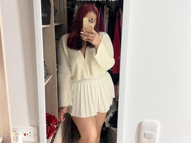 ElyRoss with red hair, white ribbed cardigan, and pleated skirt, taking a mirror selfie, showing her ass, in a room with a clothes rack and a white wall.