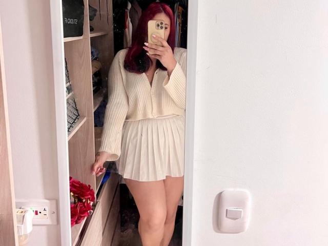 ElyRoss with red hair, covering face with phone, wearing cream ribbed top and white pleated skirt, standing in mirror selfie, showing bare thighs and butt.