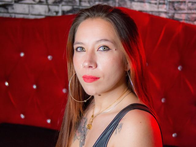 ValeFernandez has long brown hair, red lipstick, hoop earrings, and a tattoo on her chest. She's wearing a black sleeveless top, looking at the camera with a slight smile.