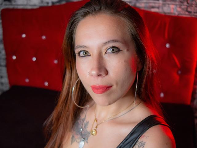 ValeFernandez has green eyes, red lipstick, and a nose ring. She wears hoop earrings, a black top, and a necklace with a pendant. Red light highlights her face.