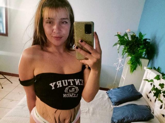 SamanthaaMiller with medium tits in a black off-shoulder top, white panties, holding a phone, standing in a room with a bed, blue wall, and green plant.