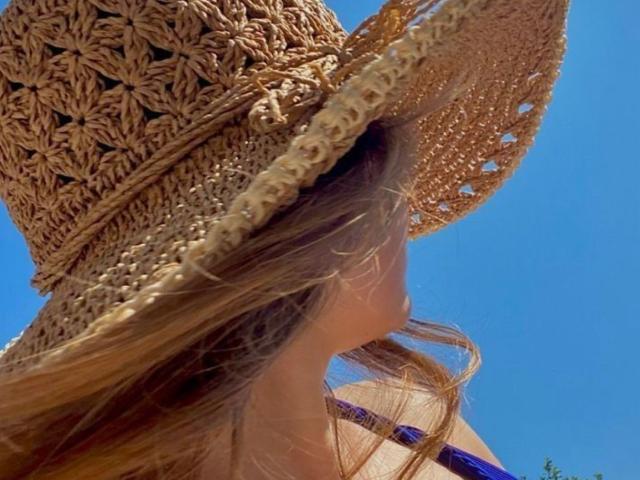 MariaViet trägt einen großen, gewobenen Strohhut mit Blumenmustern, ihr langes, braunes Haar weht im Wind, im Hintergrund ist ein klarblauer Himmel zu sehen, es ist ein sonniger Tag, der Hut verdeckt teilweise ihr Gesicht..