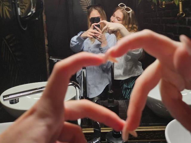 Anna et Lera prennent un selfie devant un miroir de salle de bain, leurs mains étant au premier plan, Anna portant une chemise bleue et des lunettes de soleil blanches, et tenant un téléphone..