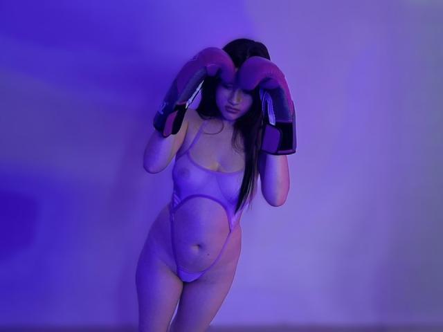 Iris Blair lleva una tanga púrpura transparente con corte alto y guantes de boxeo rosas grandes y redondos, de pie en una iluminación azul-púrpura, mostrando sus pechos y un trasero pequeño y redondo..