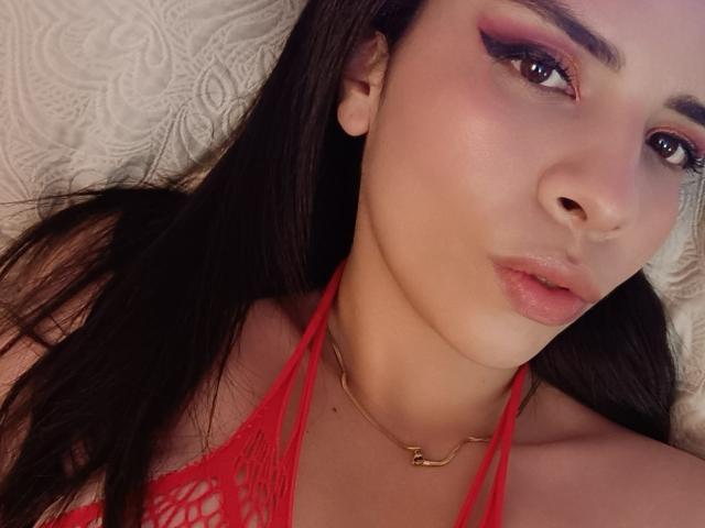 VioletaStone with pink eye makeup, red lace top, glossy lips, and a gold necklace, lying on a textured white surface, showing her face and upper chest.