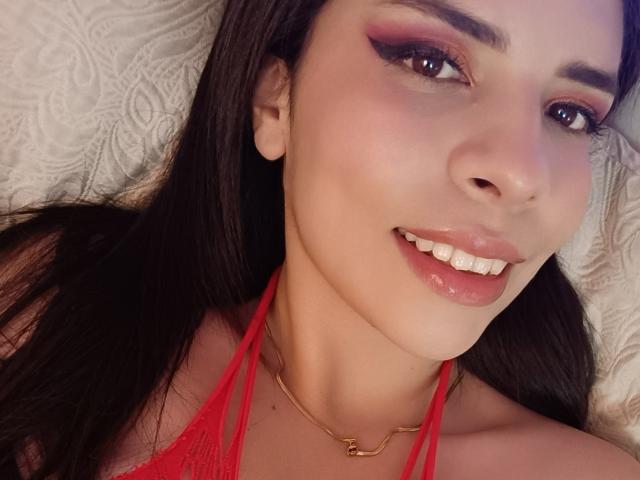 VioletaStone with dark hair, red eyeliner, and glossy pink lips, smiling, wearing a red lace halter top and gold necklace, lying on a white quilted surface.