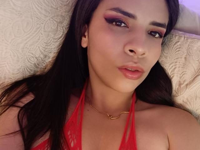 VioletaStone with long dark hair, red eyeliner, and glossy pink lips, wearing a red lace halter top, lying on a white pillow, looking at the camera.