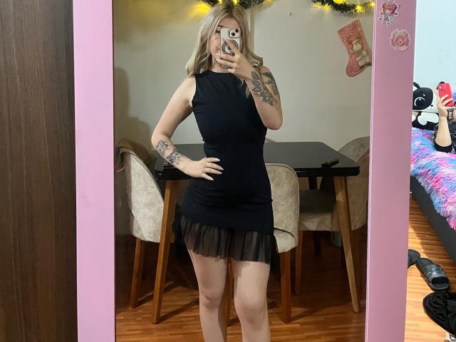 SammyViolet with blonde hair, wearing a black sleeveless dress with a flirty black frill, showing tattoos on her right arm, taking a mirror selfie, holding a phone.