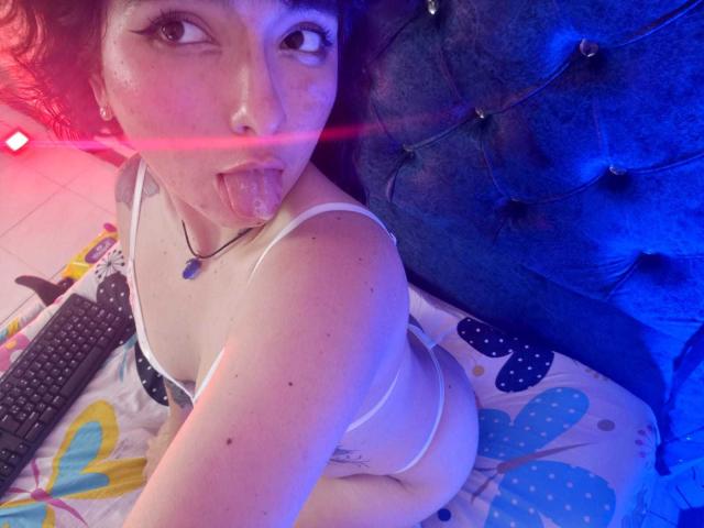 HairyValeri with dark curly hair, tongue out, wearing white bra and panties, showing ass, on blue tufted bed, in colorful patterned bedsheet, with TV remote nearby.